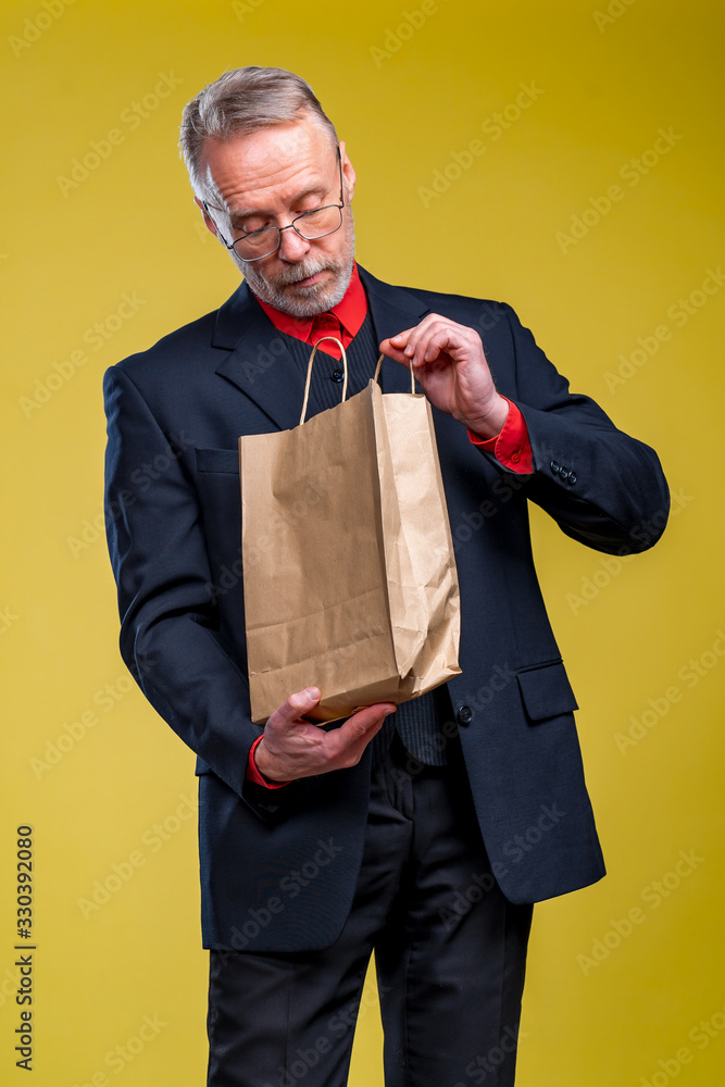 Senior man is looking at a paper bag receiving a present. Eco shopping. Man in suit on yellow backgr