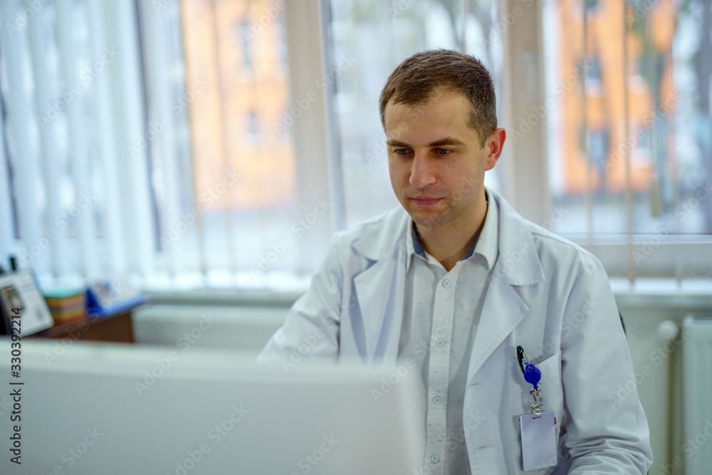 Medical office. Male doctor sitting, looking at laptop computer. Siiting at table.