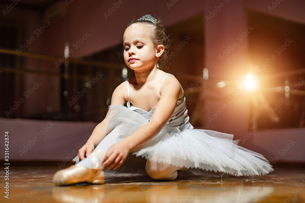 Little ballerina in white tutu sitting on floor and resting in dance studio. Pretty small girl in ba