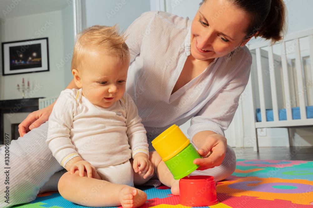 Young mother play and interact with toddler baby boy in the nursery sitting on the floor holding pla