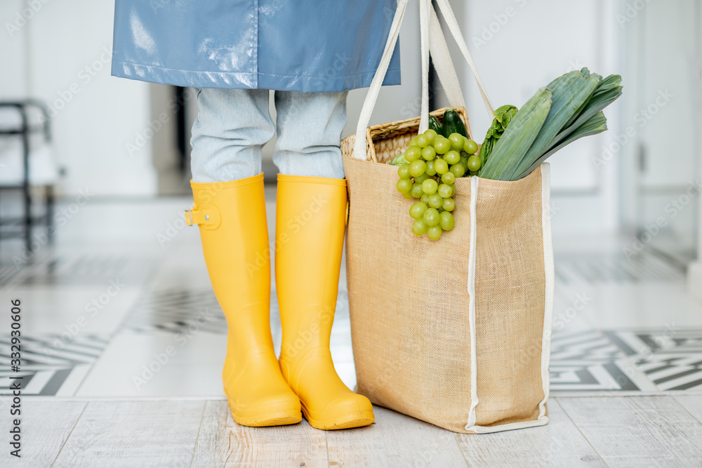 穿着黄色橡胶靴和雨衣的女人站在走廊里，手里拿着装满新鲜食物的购物袋