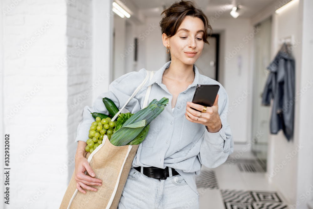 一个年轻开朗的女人的画像，手里拿着手机和装满新鲜食物的购物袋
