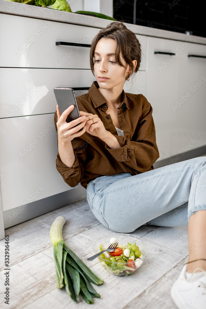年轻女子拿着手机坐在厨房地板上，一边吃沙拉，一边在家放松。健康的ea