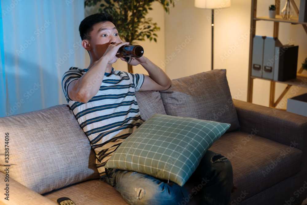 Young asian chinese man watching television drinking bottle of beer in late evening time. handsome g