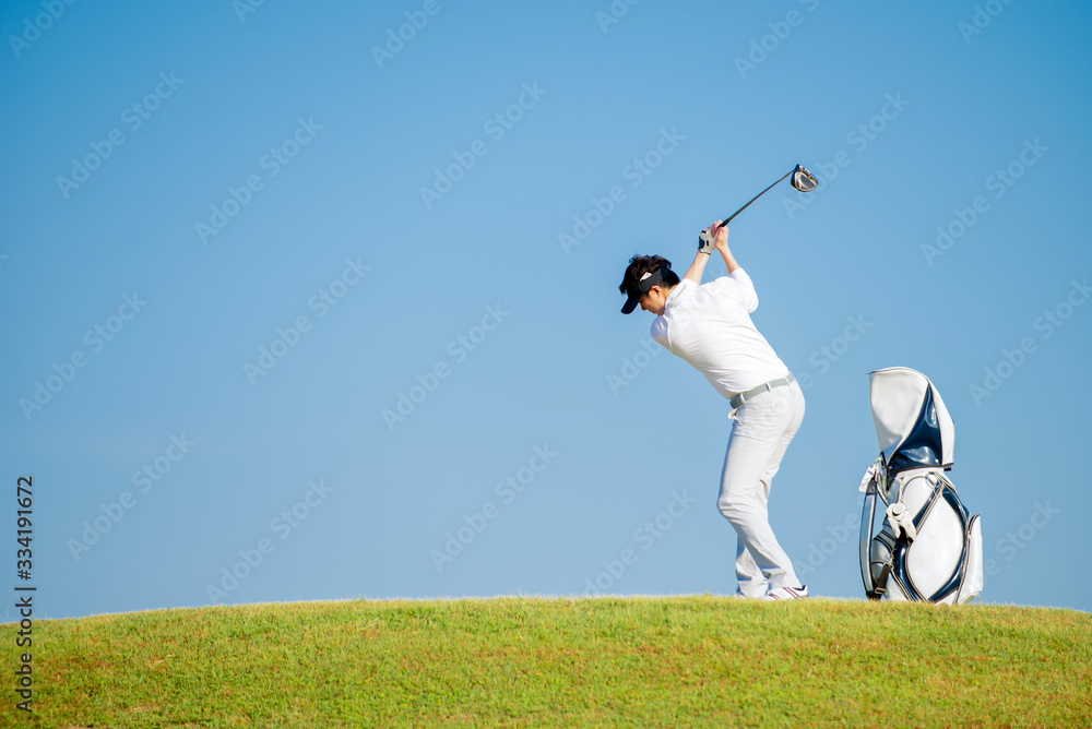 亚洲男子高尔夫球手拿着高尔夫球袋站在斜坡上，在高尔夫球场的蓝天背景下击球