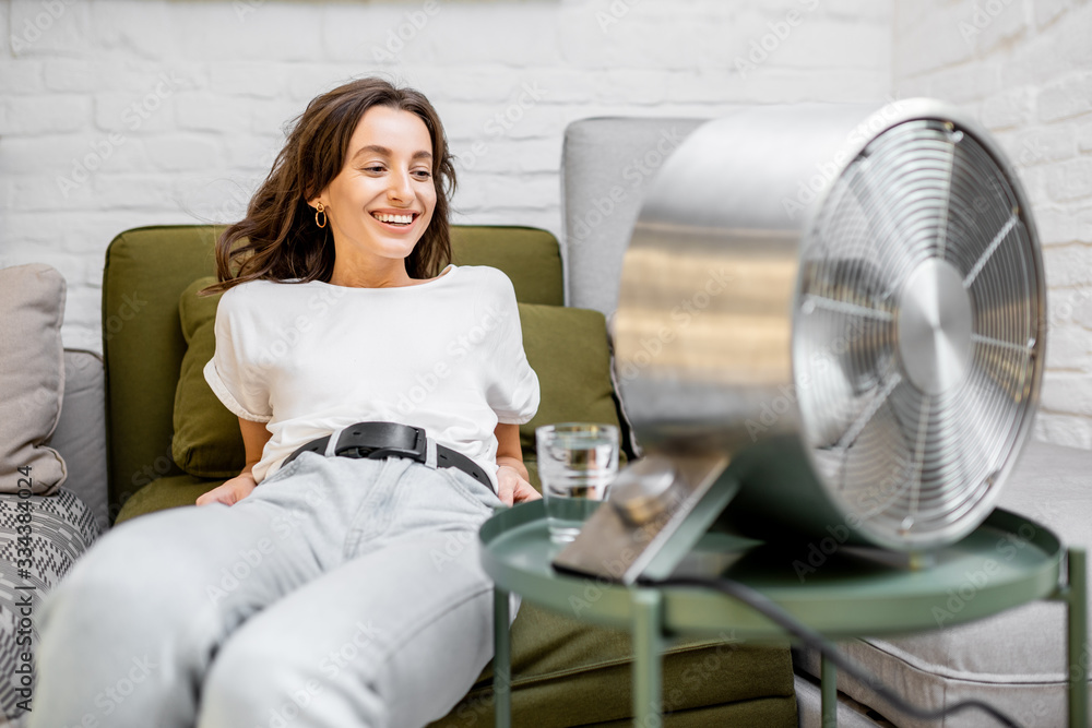 Young woman feeling hot, enjoying the flow of air from the fan, sitting on the couch with a glass of