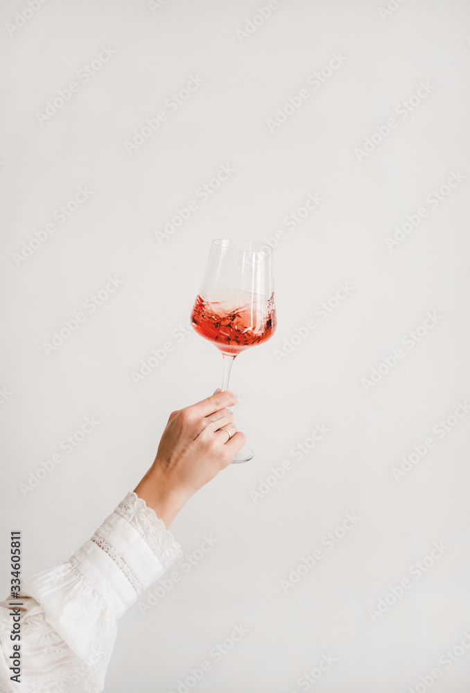 Womans hand in white shirt holding and turning glass of rose wine over white wall background. Wine s