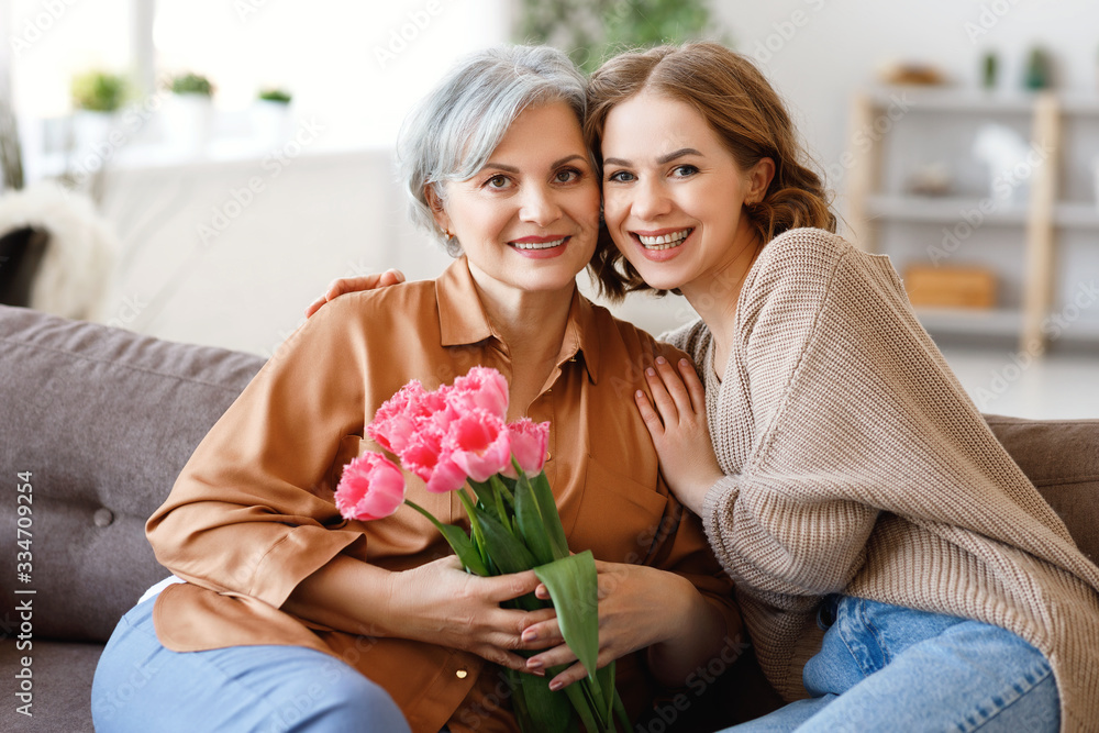Happy woman hugging aged mother with flowers.