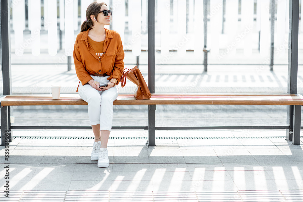阳光明媚的一天，快乐时尚的女人带着手机和包坐在现代公共交通站