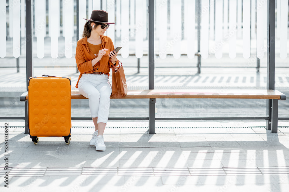 年轻的女性旅行者提着行李箱坐在交通站等电车或公交车。Di