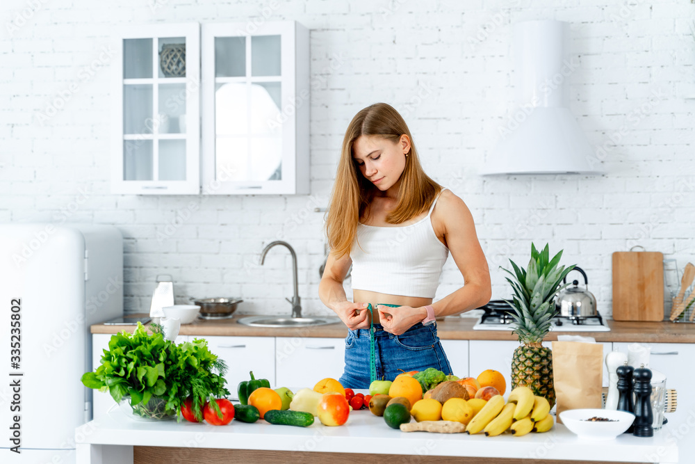 Young slim girl with measuring tape. Womn standing in the modern kitchen. Fruits and vegetables on t