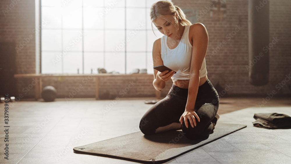Beautiful and Young Girl Uses Smartphone App During Her Exercise on Fitness Mat. Gorgeous and Athlet