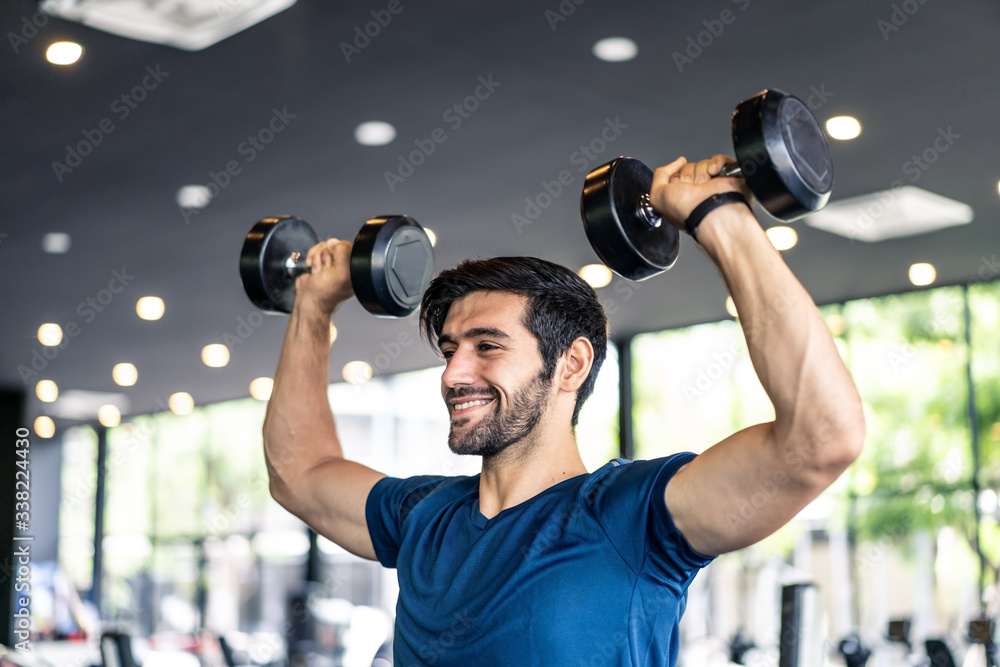 年轻健康的高加索英俊男子在健身房或健身俱乐部双手举哑铃。男性在
