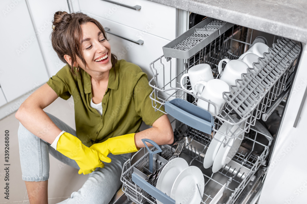 Portrait of a happy housewife sitting near the dishwasher with clean dishes on the kitchen at home. 