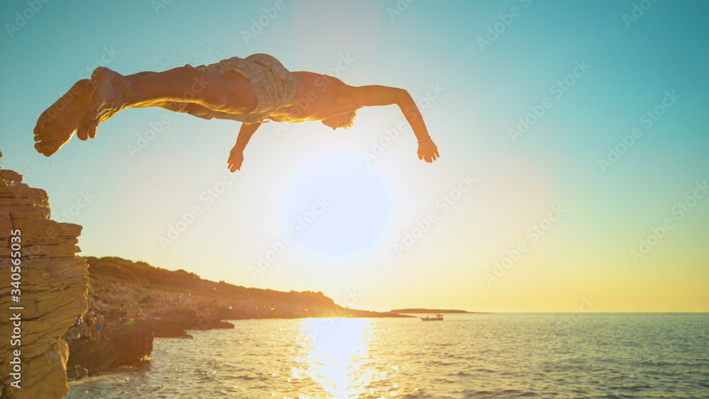 CLOSE UP: Golden sunset shines on rock diver jumping off a cliff and into sea.