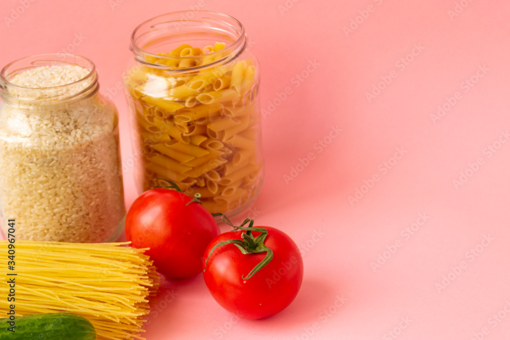 Noodles, rice, pasta in glass jars stand on a pink background. Nearby are red tomatoes and green cuc