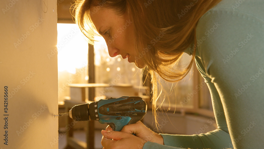 CLOSE UP: Young woman uses a power drill to screw in a bolt into a wall panel.