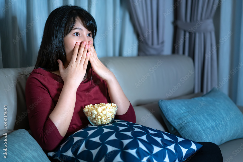 Young asian woman watching television suspense movie or news looking shocked and excited eating popc