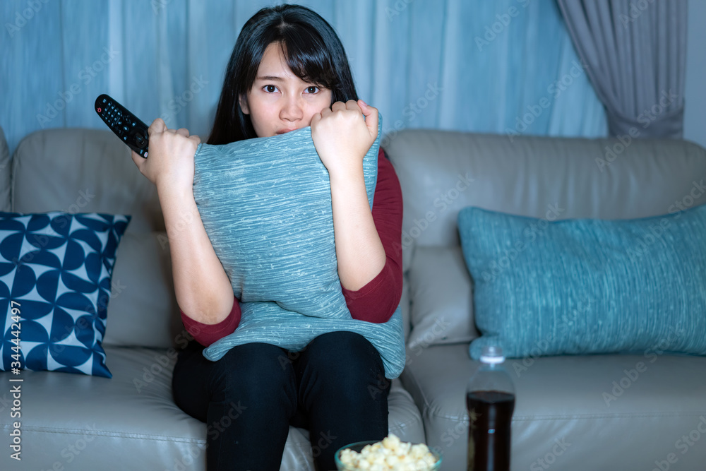 年轻的亚洲女性在看电视悬疑电影或新闻时看起来既震惊又兴奋地吃着爆米花