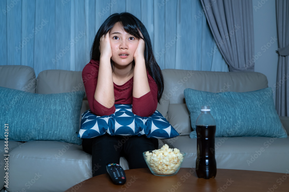 Young asian woman watching television suspense movie or news looking shocked and excited eating popc