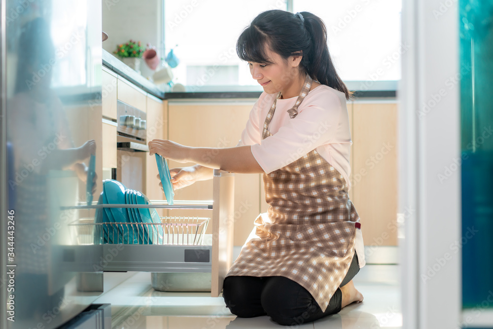 Attractive young Asian woman loading the dishwasher into cupboards at kitchen while doing cleaning a