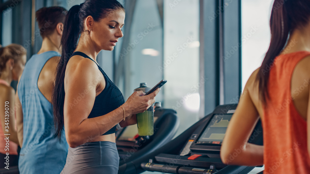 健身房里美丽的运动型女性在跑步机上跑步，使用智能手机并饮用蛋白质Su
