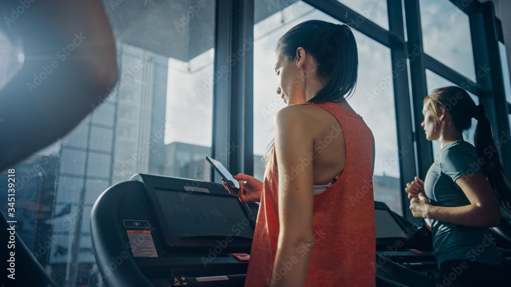 健身房里漂亮的运动女，在跑步机上跑步，使用智能手机。背景无线