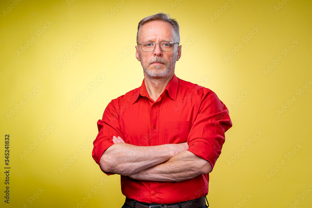 Aged bearded fit man is standing with crossed hands isolated against yellow background. Red shirt. M