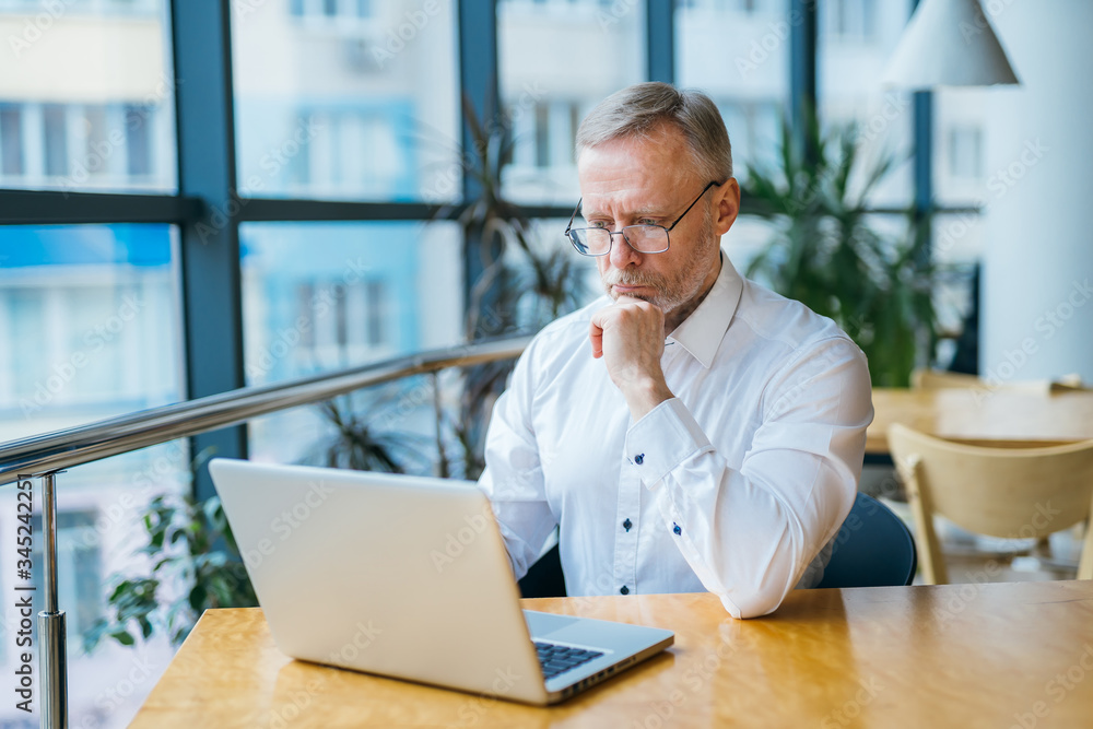 Thoughtful middle aged businessman in white shirt. Man sitting with a laptop near the window working
