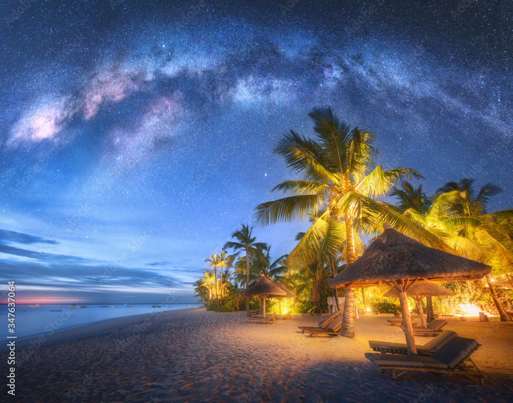 银河系在沙滩上，夏天晚上有棕榈树、日光浴床和雨伞。Landsca