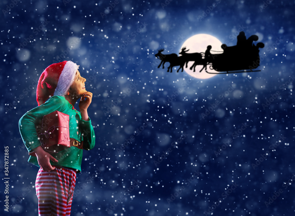 带着新年装饰品的快乐体贴的小男孩看着圣诞老人驯鹿雪橇的轮廓