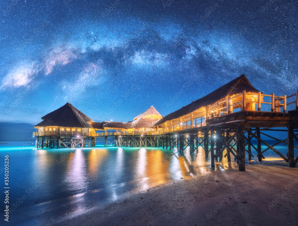 Milky Way over wooden bungalow on the water in summer starry night. Landscape with hotel on the sea,