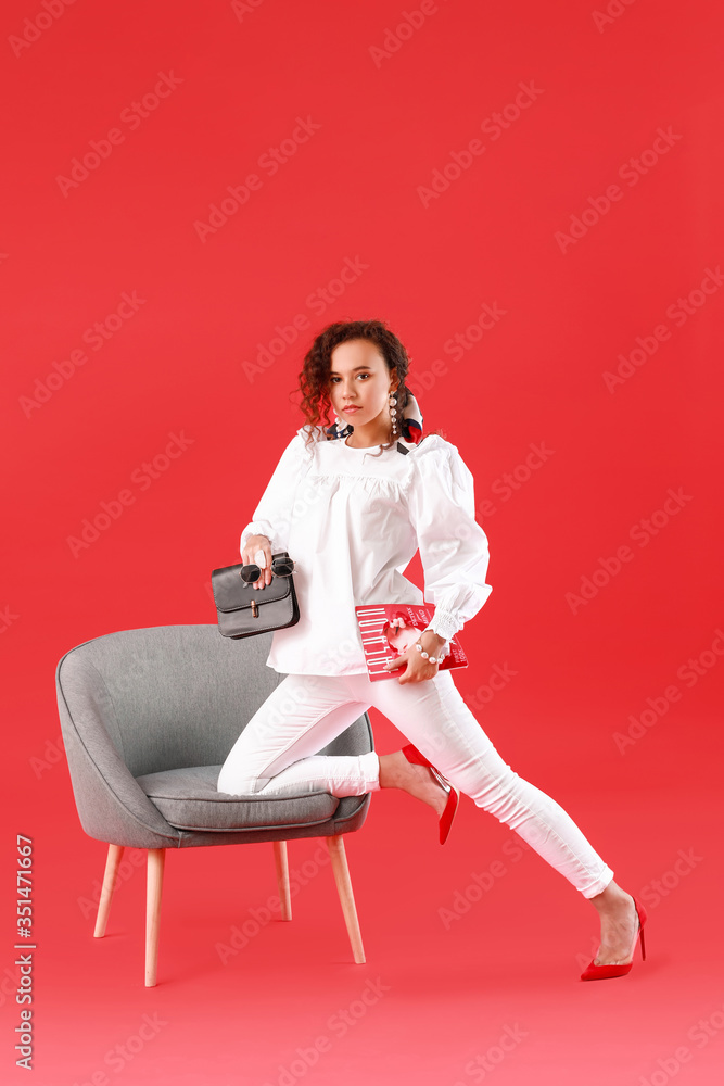 美丽的年轻非裔美国女性，时尚的包包和杂志放在扶手椅附近的彩色背景上