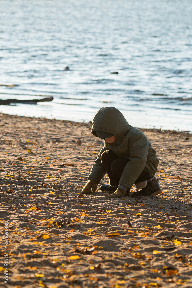 一个穿着带风帽夹克的男孩坐在海边，在沙滩上玩耍，背后是海浪