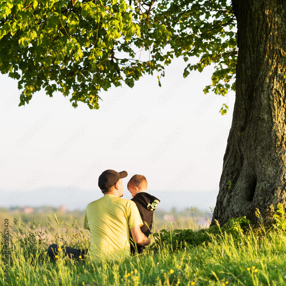爸爸和儿子在春天的草地上坐在高高的草地上的树下。带着孩子旅行的概念