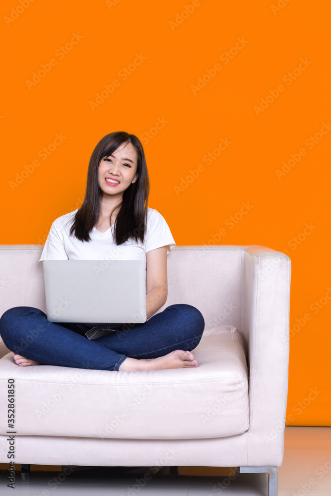 Asian woman smiling and working from home use laptop computer while sitting on sofa over isolate yel