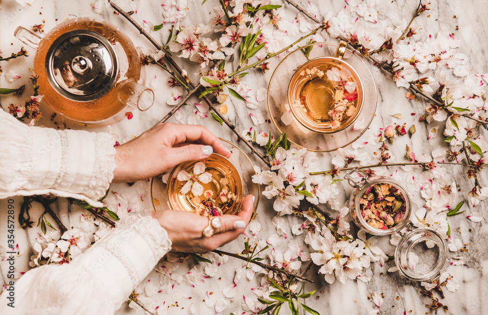女性双手平放，手里拿着一杯热红茶、茶壶、罐子里的干玫瑰和春天的bl