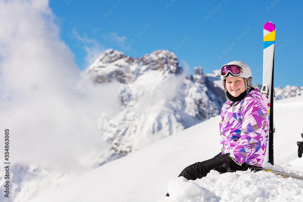 Happy girl sit in the mountain with ski over high peaks and clouds in snow look at camera smiling