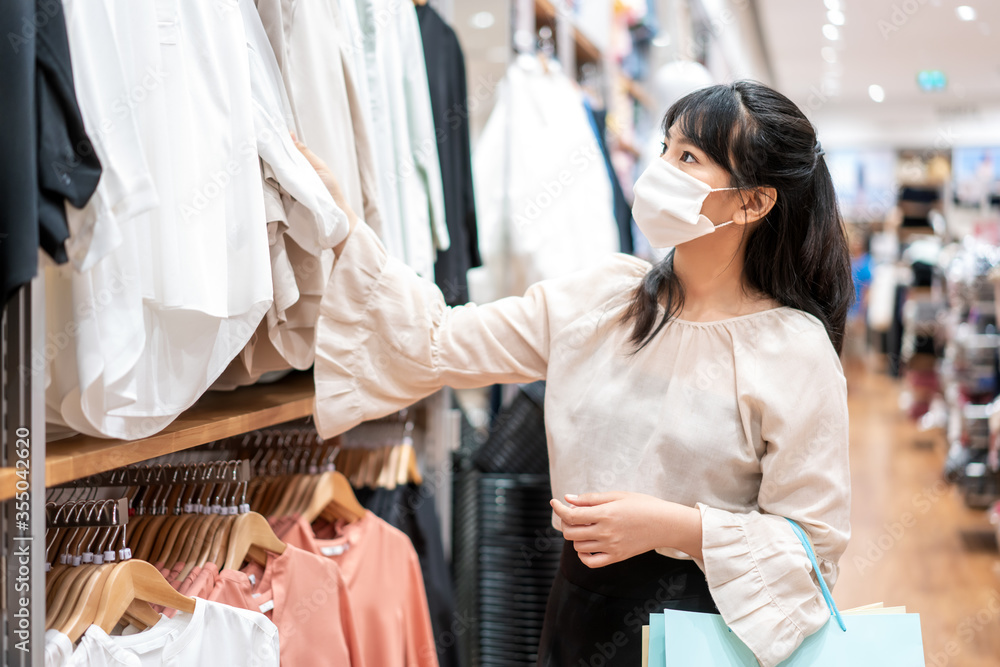 Asian woman wearing mask over her face while choosing shirt at shopping mall with shopping bag for h