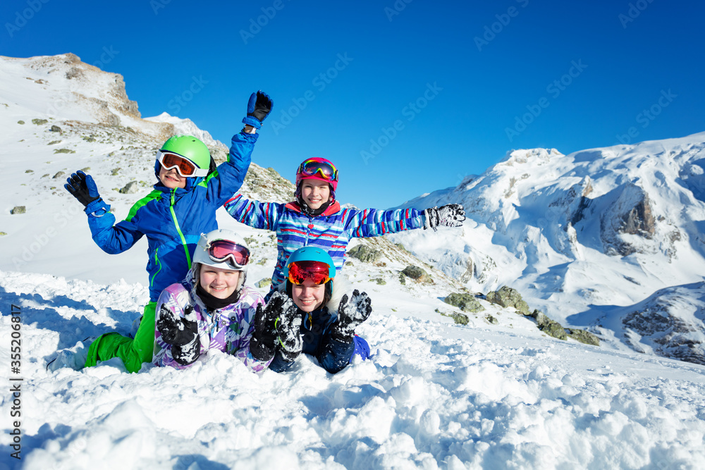 四个快乐的孩子一起躺在山顶的雪地里，穿着滑雪服微笑着挥手