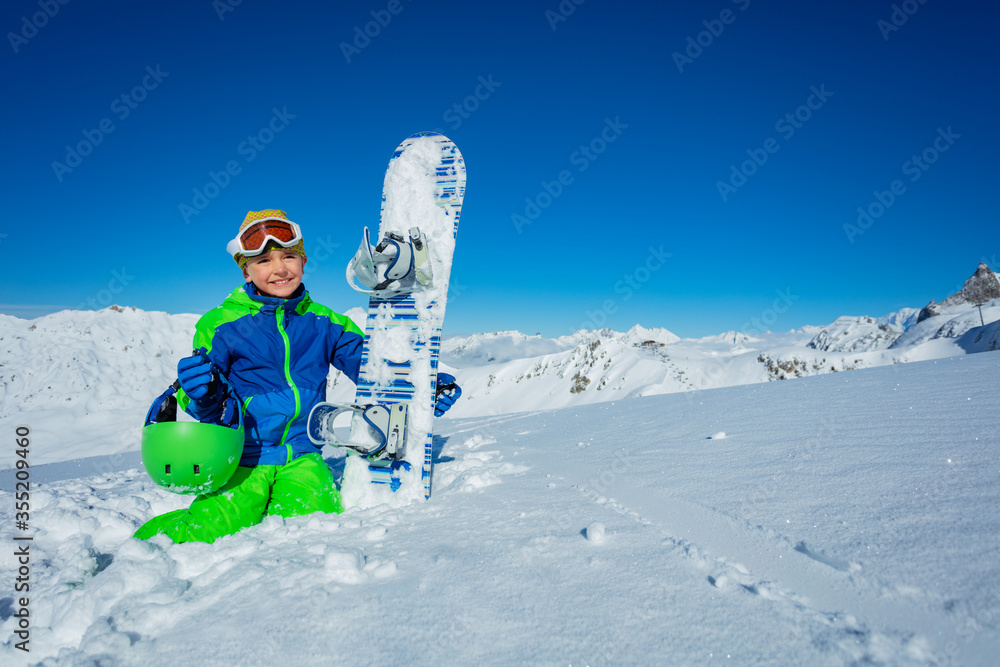 一个男孩的肖像，他在雪地里拿着滑雪板，戴着休闲帽，在良好的天气下俯瞰山脉全景