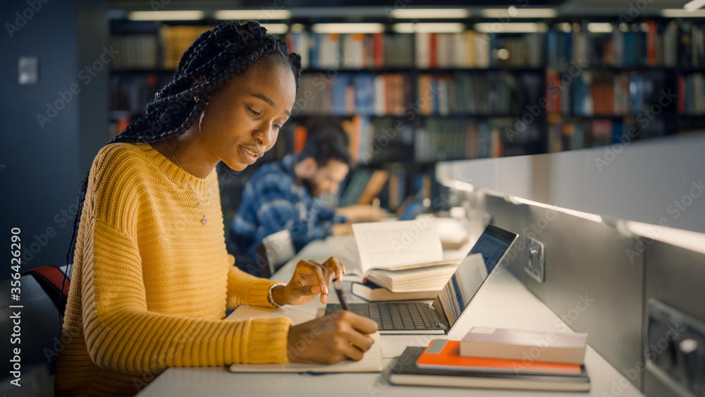 大学图书馆：天才美丽的黑人女孩坐在桌子旁，用笔记本电脑，为t写笔记