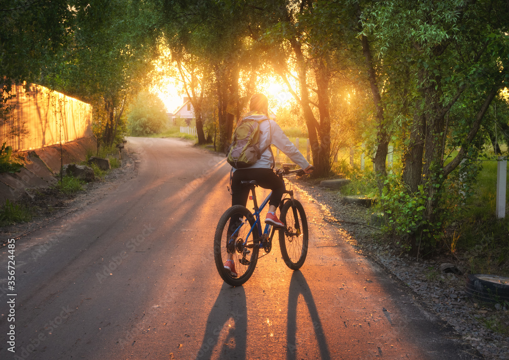 夏天日落时，一个女人骑着自行车在乡村公路上。五颜六色的风景带着运动女孩的wi