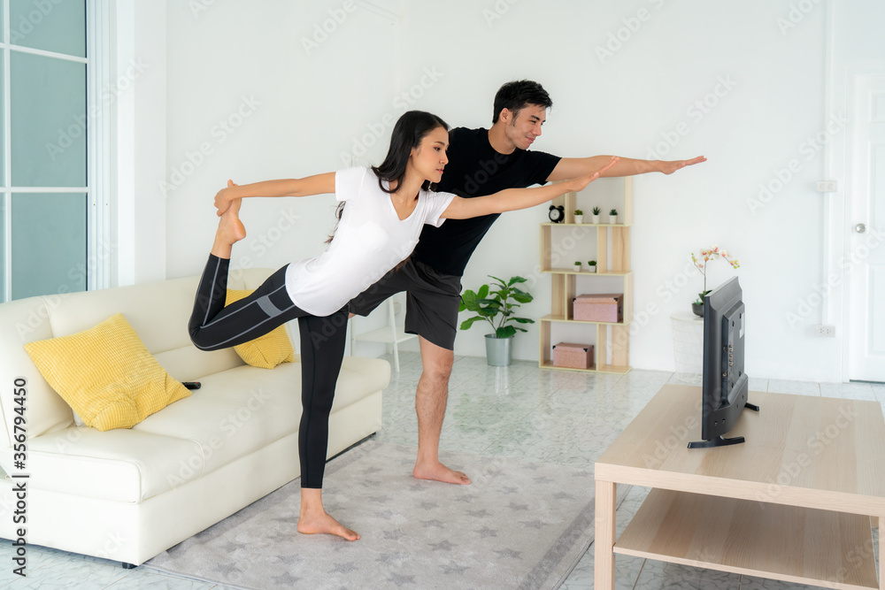 Young Asian couple doing yoga practice together and looking TV at home, man and woman working out to