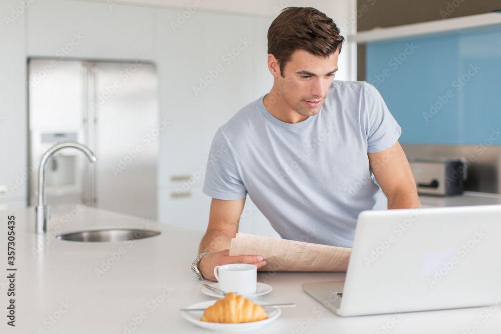 在厨房柜台使用笔记本电脑的男报，前景是咖啡杯羊角面包