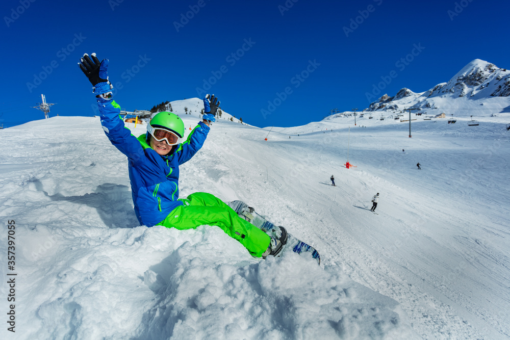 带着滑雪板的小男孩戴着口罩，戴着滑雪帽，双手高举，坐在雪地上