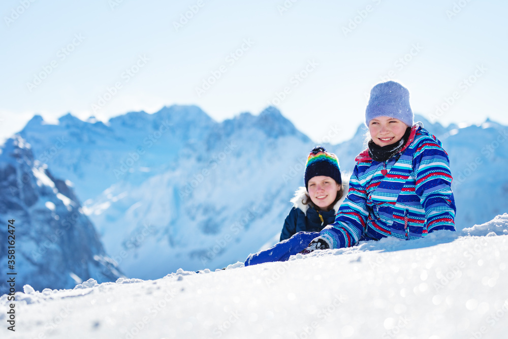 两个快乐微笑的女孩戴着冬天的帽子，穿着高山顶峰的外套，躺在雪地里