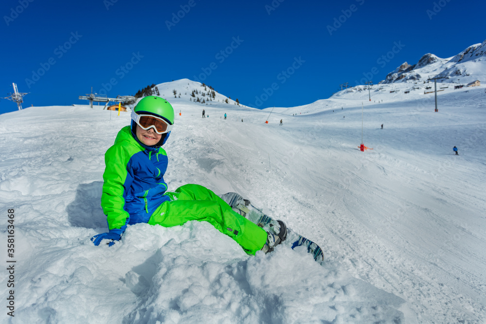 带着滑雪板的小男孩戴着口罩，戴着滑雪帽，从侧面坐在雪地上微笑