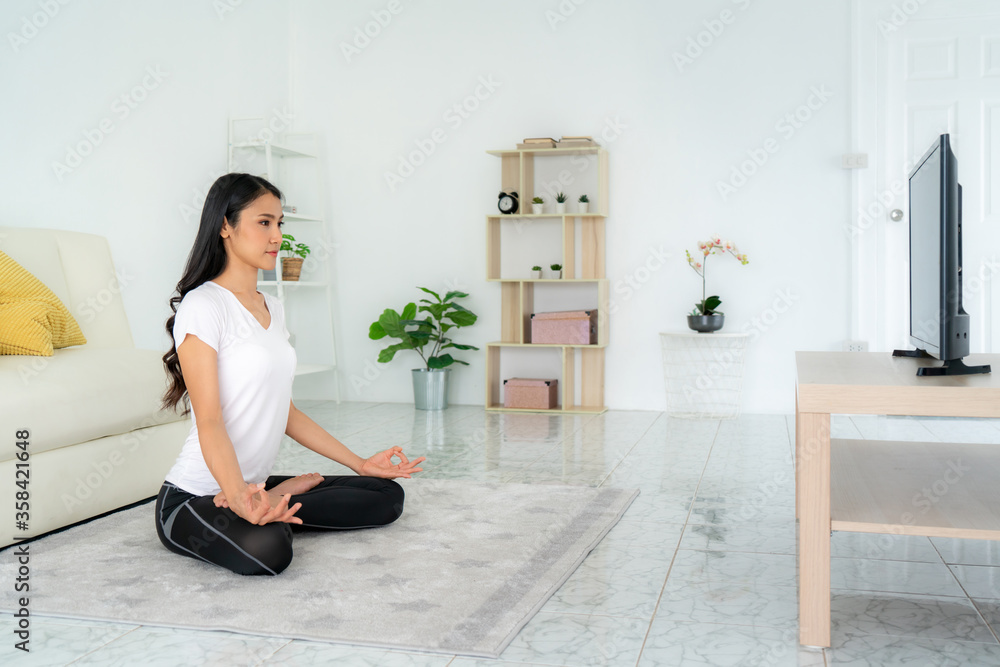 Beautiful young Asian woman in sports clothing doing yoga while relaxing in living room at home to g