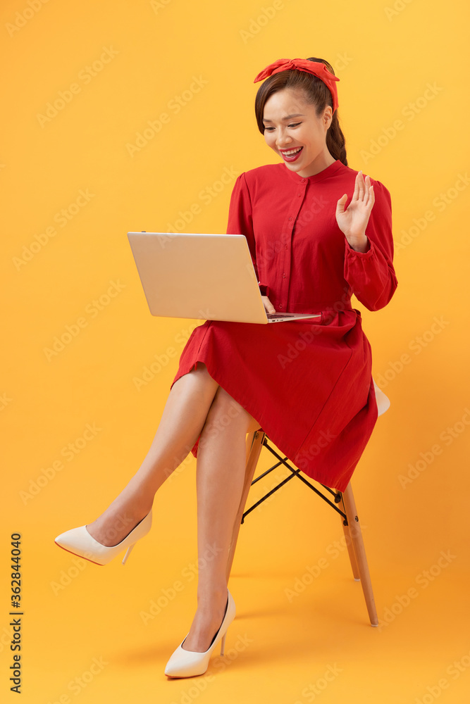 快乐的年轻亚洲女性在橙色背景的扶手椅上使用笔记本电脑。